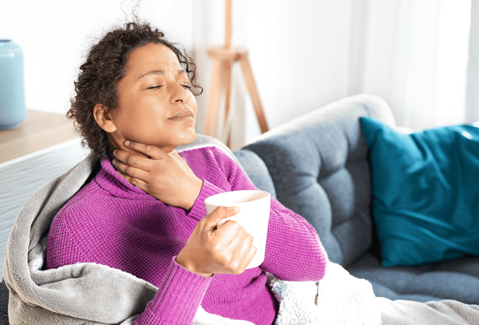 lady sat on couch with cup of tea holding sore throat