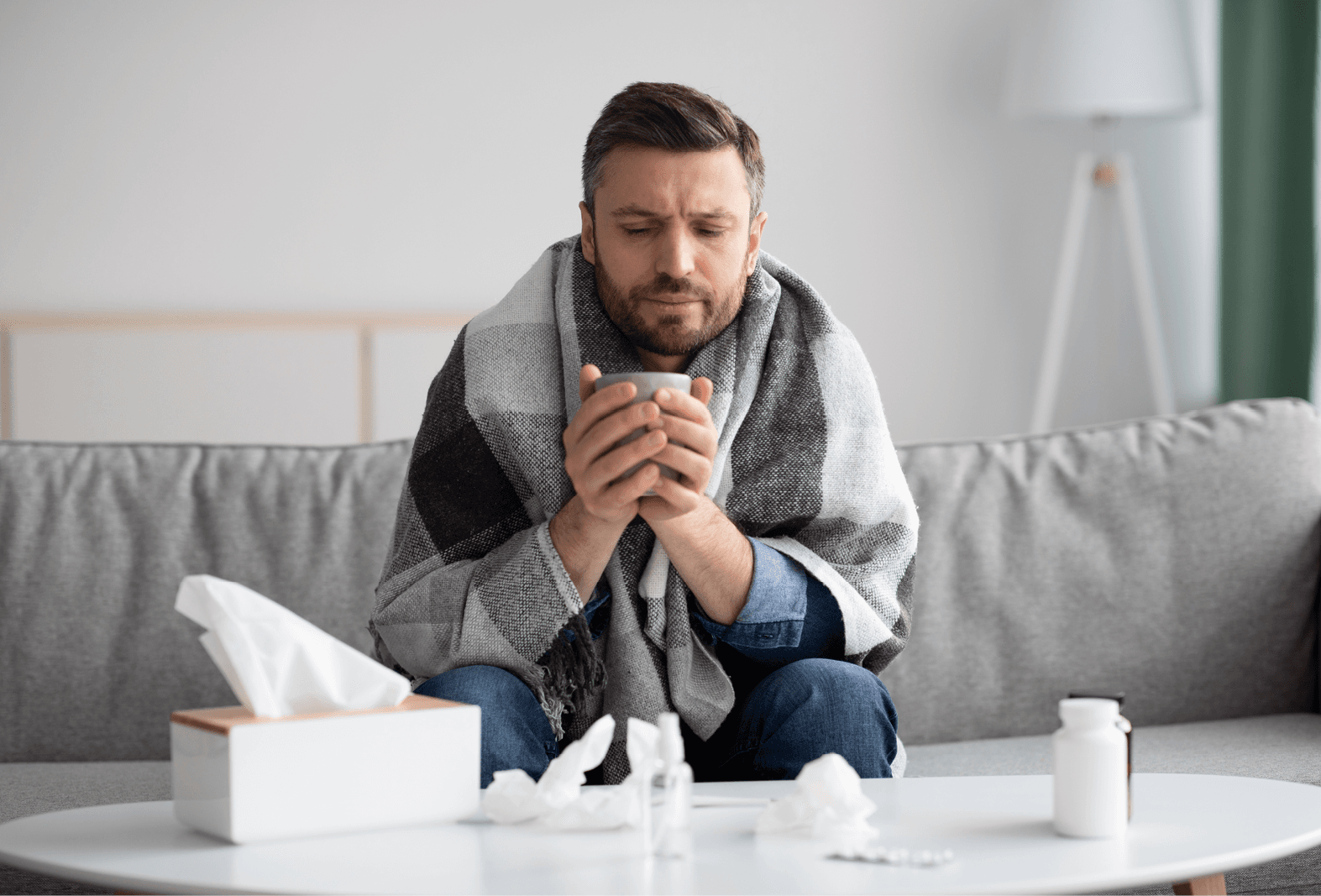 man sat on couch, wrapped in blanket, with hot cup and tissues around